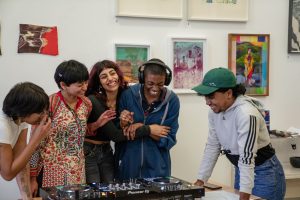 Group of young people embrace around DJ decks. They are stood in front of colourful framed artwork.