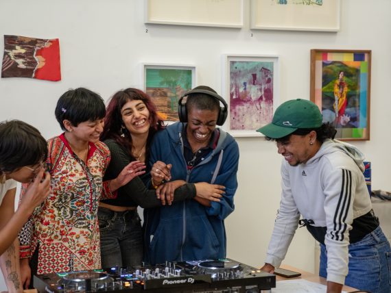 Group of young people embrace around DJ decks. They are stood in front of colourful framed artwork.