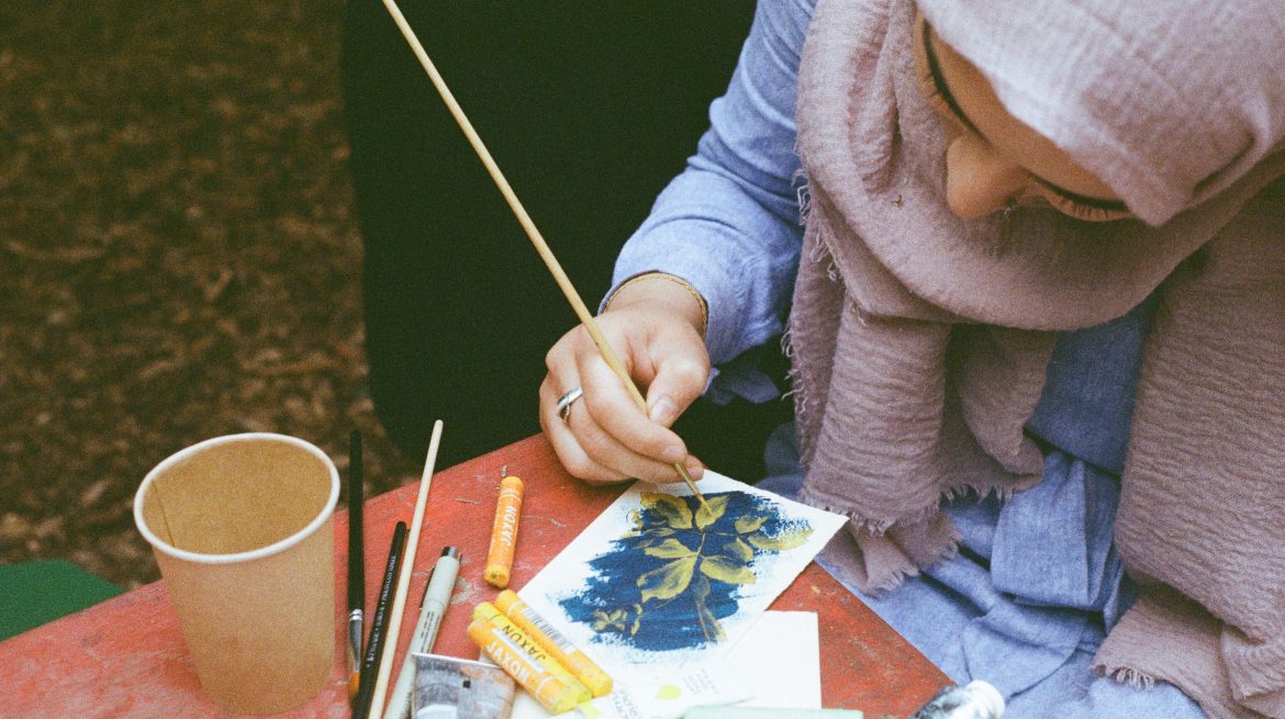 Film photograph of young woman from above painting a yellow flower onto a blue coloured piece of paper