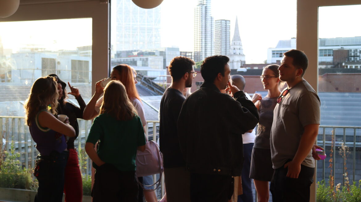 A group of young people stand in a group by an open window talking as the sun sets