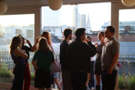 A group of young people stand in a group by an open window talking as the sun sets