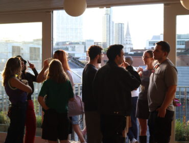 A group of young people stand in a group by an open window talking as the sun sets